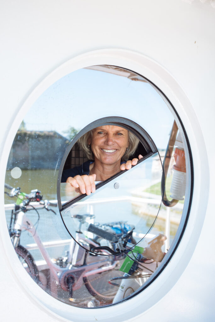 Lächelnde Frau durch ein Bullauge gesehen, während sie ihr Fahrrad auf einem Boot arrangiert