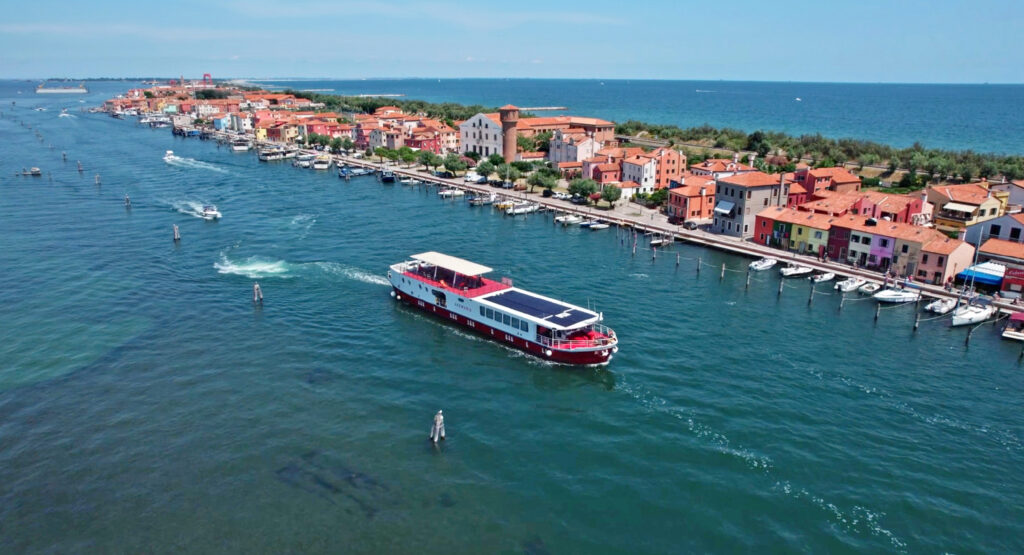 Das rot-weiße Boot Girolibero fährt auf der venezianischen Lagune mit einem Dorf aus bunten Häusern im Hintergrund
