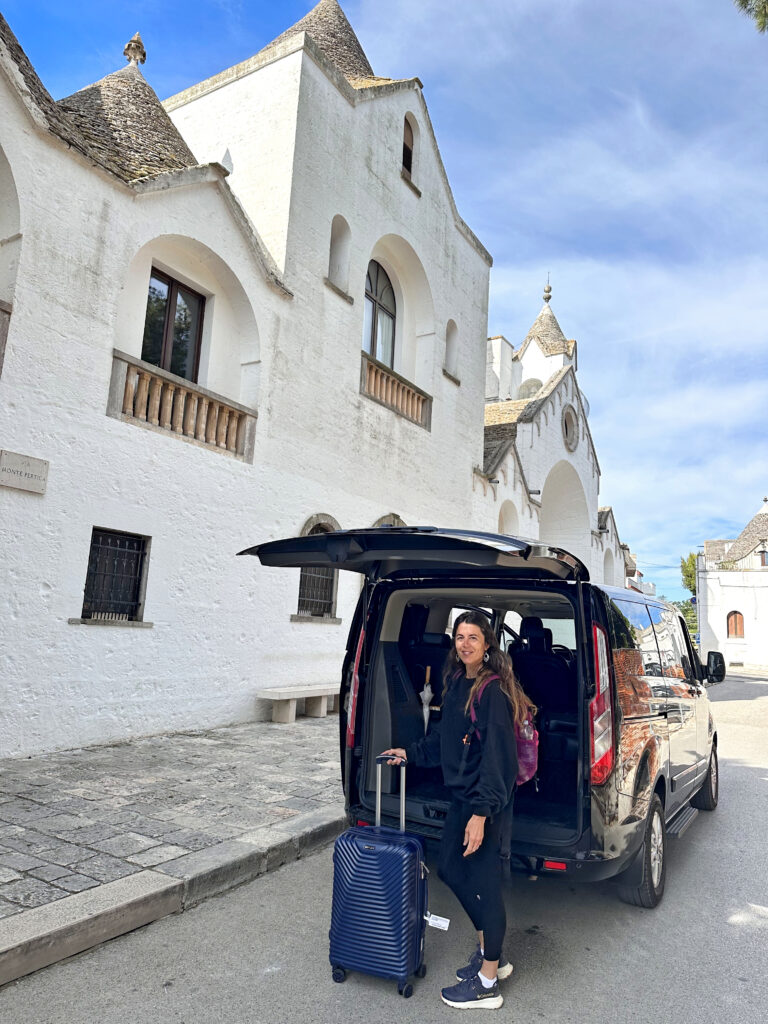 Menschen laden Gepäck in einen Lieferwagen auf einer Kopfsteinpflasterstraße neben weißen Häusern.